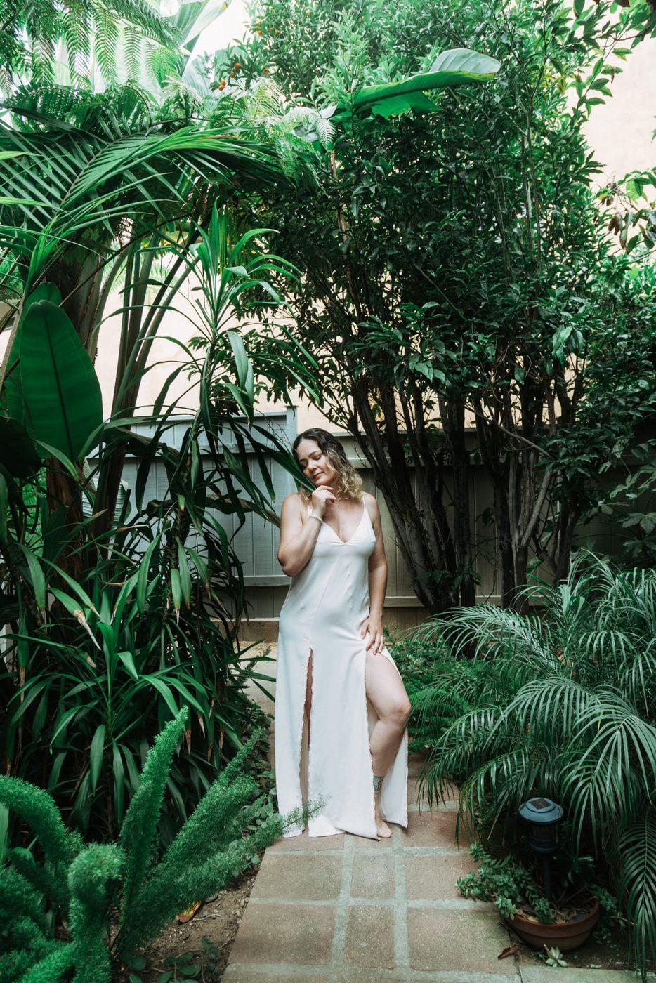 Simone Dib standing in a garden path, head tilted, in a white dress